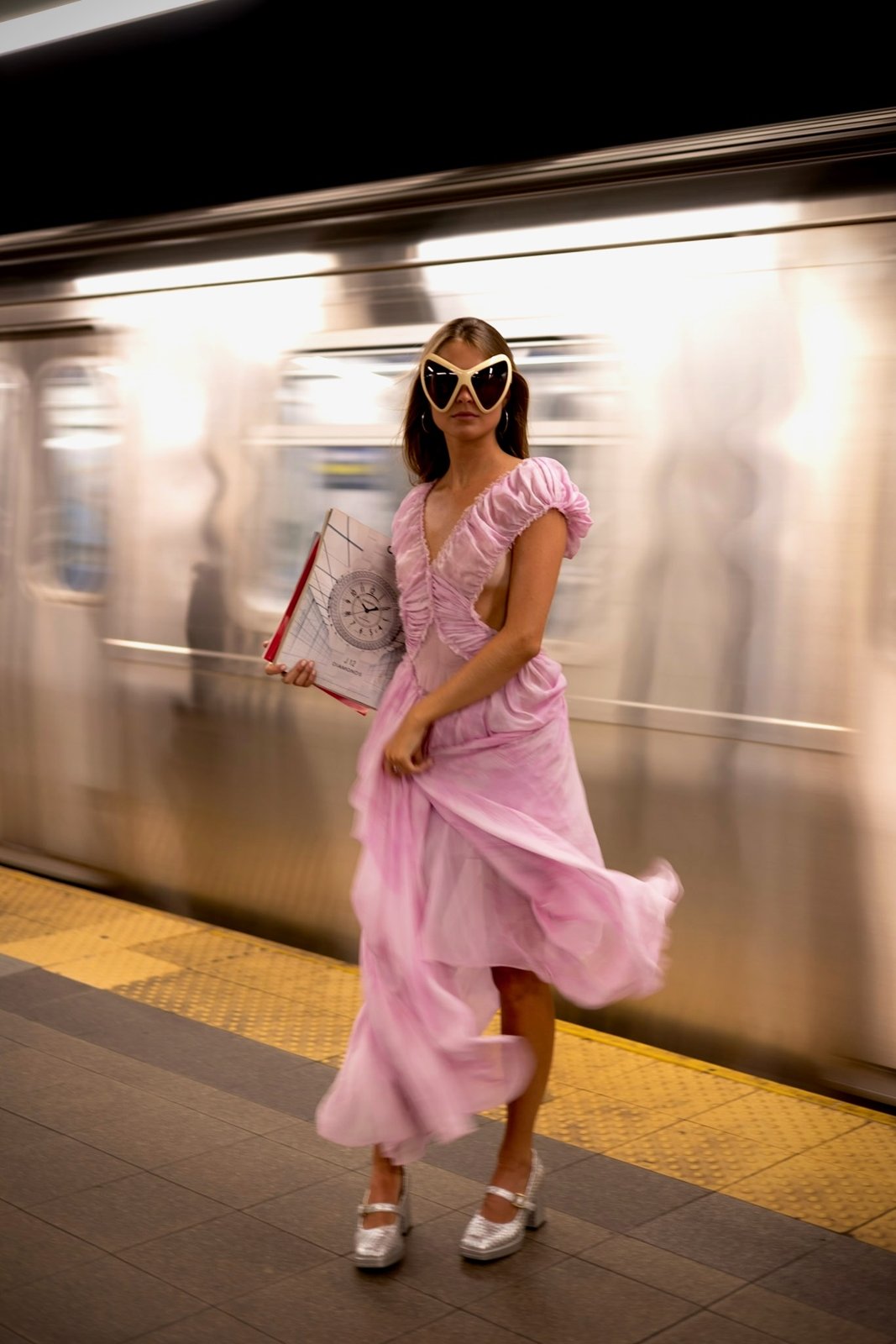 A woman in a pink dress and oversized sunglasses on a subway platform, from The Play Issue